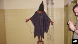Ova fotografija iz 2003. koju je dobio AP prikazuje neidentifikovanog pritvorenika kako stoji na kutiji sa torbom na glavi i prikačenim žicama u zatvoru Abu Ghraib u Bagdadu u Iraku.