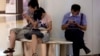 FILE - People sit on a bench inside a shopping mall using their mobile devices, in Beijing, China, Aug. 19, 2013. 