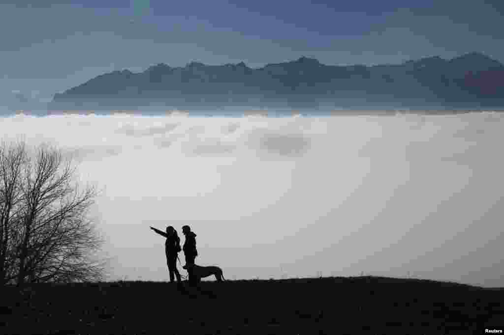 A couple walking their dog look at a sea of fog over Lake Leman at the Tour de Gourze near Lausanne, Switzerland.