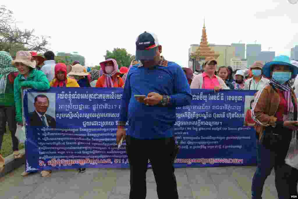 បុរសសន្តិសុខស្លៀកពាក់ស៊ីវិលម្នាក់ កំពុងឈរនៅខាងមុខបដាដែលក្រុមកម្មករកម្មការិនីរាប់រយនាក់ មកពីរោងចក្រ W&D Cambodia Co., LTD សុំអន្តរាគមន៍ពីនាយករដ្ឋមន្ត្រី ហ៊ុន សែន ភ្នំពេញ នៅថ្ងៃទី១៨ ខែមីនា ឆ្នាំ២០១៩។ (កាន់ វិច្ឆិកា/VOA)